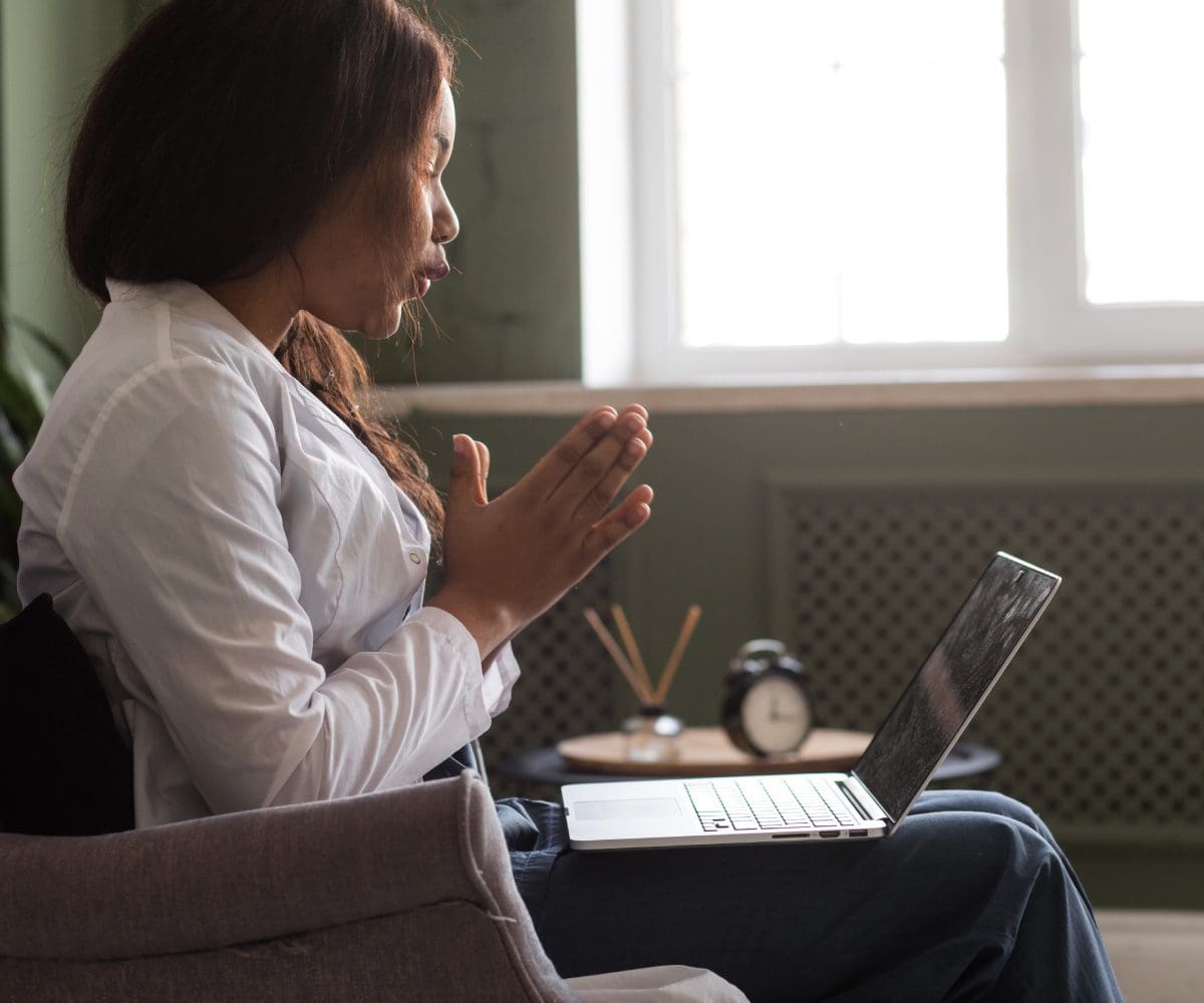 A woman sits by a window, holding a mug and looking at her laptop.